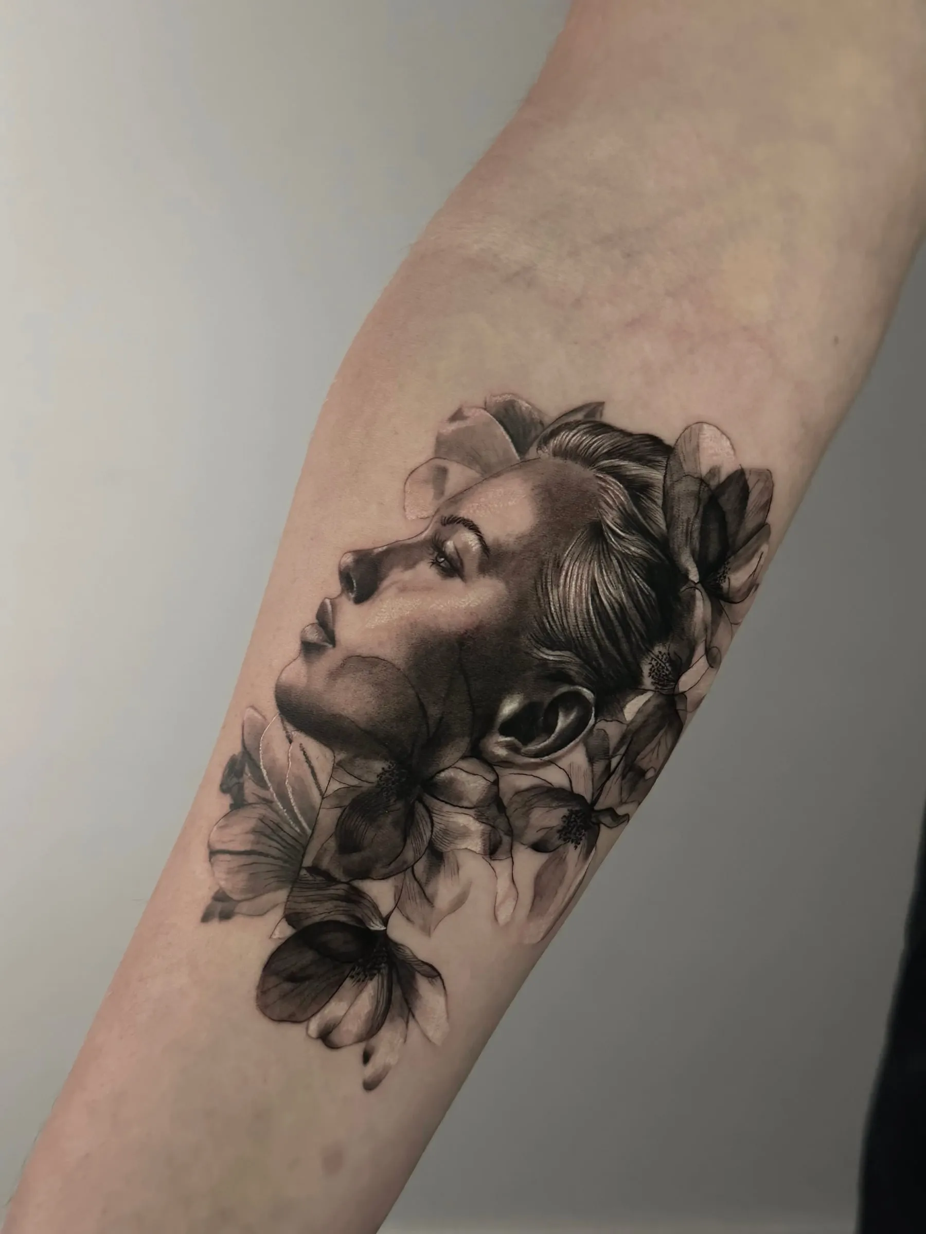 Retrato realista en negro y gris de un perfil de mujer rodeado de flores, en el antebrazo